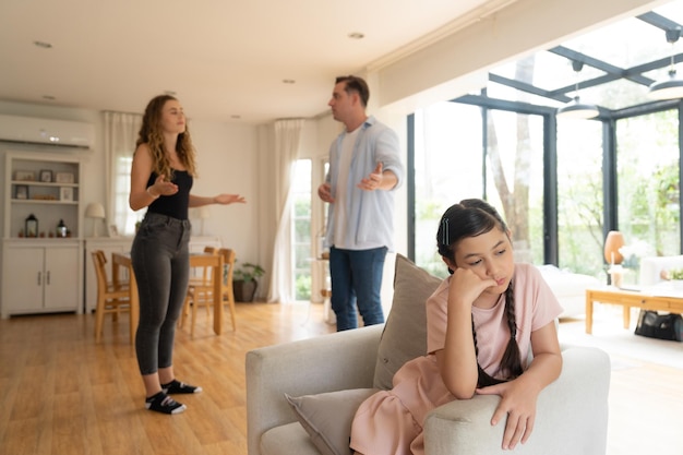 Annoyed and unhappy young girl sitting on sofa trapped in middle of tension by her parent argument in living room Unhealthy domestic lifestyle and traumatic childhood develop to depressionSynchronos