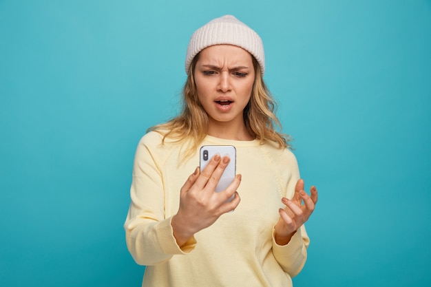 Annoyed young girl wearing winter hat holding and looking at mobile phone keeping hand in air 