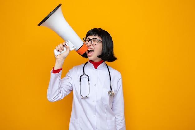 Annoyed young pretty caucasian girl with optical glasses in doctor uniform with stethoscope holding and shouting into loudspeaker standing with closed eyes isolated on orange wall