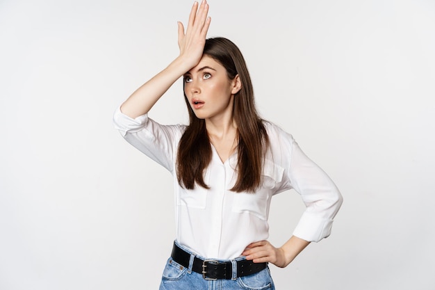 Annoyed young woman facepalm, slap forehead and roll eyes bothered, standing in white shirt and jeans over white background.