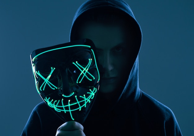 Anonymous man in black hoodie hiding his face behind a neon mask