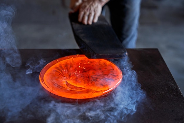 Anonymous professional male master working with hot orange glass carefully at dark table with smoke in factory