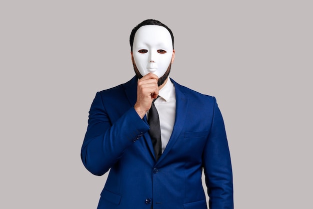 Anonymous unknown businessman covering her face with white mask, hiding her real personality, anonymity, wearing official style suit. Indoor studio shot isolated on gray background.