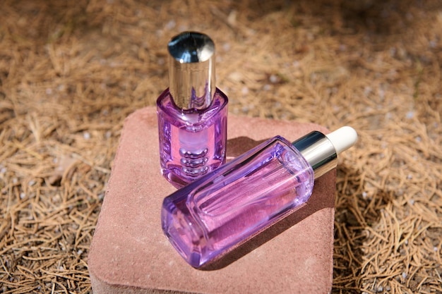 Antiaging serum in purple jars with dropper on a pink stone with sunlight on the background of fallen dry pine needles