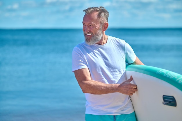 Anticipation. Gray-haired bearded sporty man with surf boat smiling looking away standing on seashore on summer day