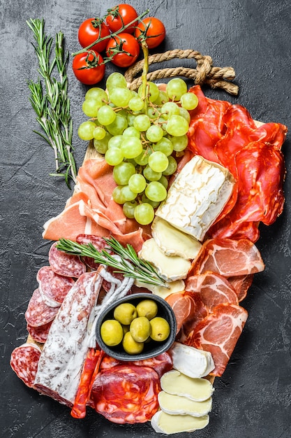 Antipasto platter cold meat with grapes, prosciutto, slices ham, beef jerky, chorizo salami, fuet, Camembert and goat cheese.  Black surface. Top view