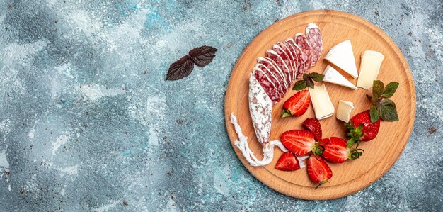 Antipasto sliced Spanish Fuet salami wurst, Camembert cheese, strawberries and glass rose wine on blue background. Long banner format. top view.