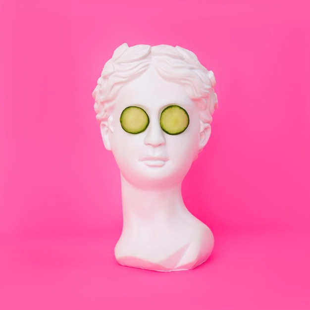 Antique female statue's head with cucumber slices on her eyes isolated on a pink background