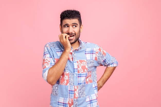 Anxiety disorder. Portrait of stressed out, worried man in blue casual shirt biting nails, nervous about troubles, panicking and looking scared. Indoor studio shot isolated on pink background.