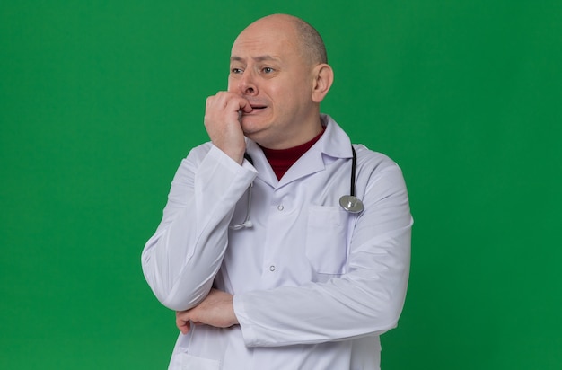 Anxious adult slavic man in doctor uniform with stethoscope biting his nails looking at side 