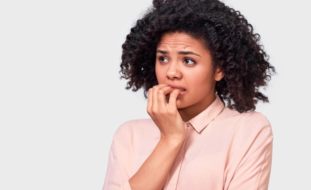 Anxious fearfull African American young woman keeps hand near mouth feels frightened and scared wearing in beige shirt standing against white studio wall Facial expressions emotions and feelings