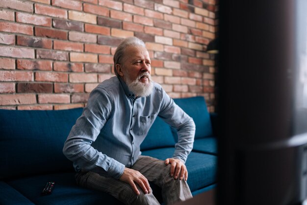 Photo anxious grayhaired mature adult male sit relax on couch in living room watch tv bearded senior older man rest on sofa at home feel worried about news