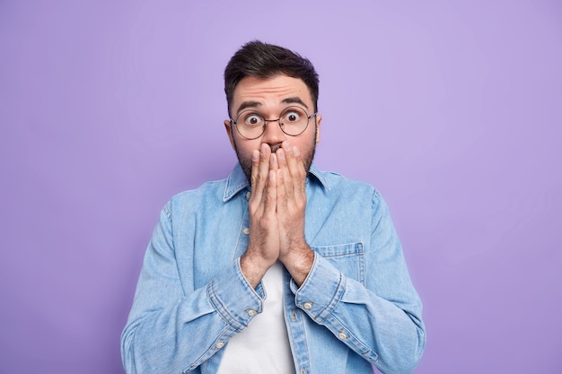 Photo anxious shocked speechless adult man covers mouth afraids of somethig terrible hears awful scary news dressed in denim shirt round spectacles