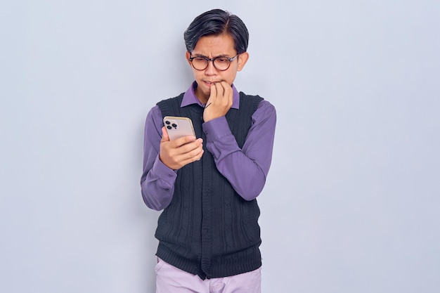 Anxious young Asian man in casual shirt using smartphone and biting nail isolated on white background