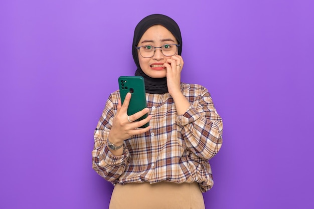 Anxious young Asian woman in plaid shirt holding a mobile phone biting nails with worried expression isolated on purple background