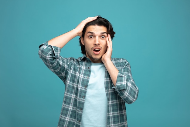 anxious young handsome man keeping hand on head and on face looking at camera isolated on blue background