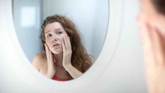 Anxious young woman look in the mirror worried about wrinkle or acne on unhealthy skin upset unhappy millennial female examine squeeze pimple on face in the bedroom cosmetology skincare concept