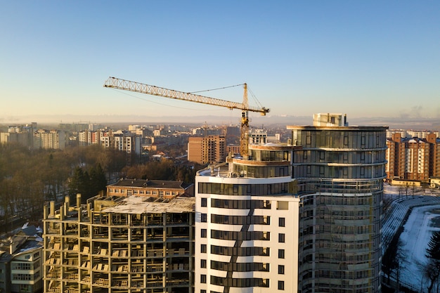 Apartment or office tall building under construction, top view. Tower crane on bright blue sky copy space, city landscape stretching to horizon. Drone aerial photography.