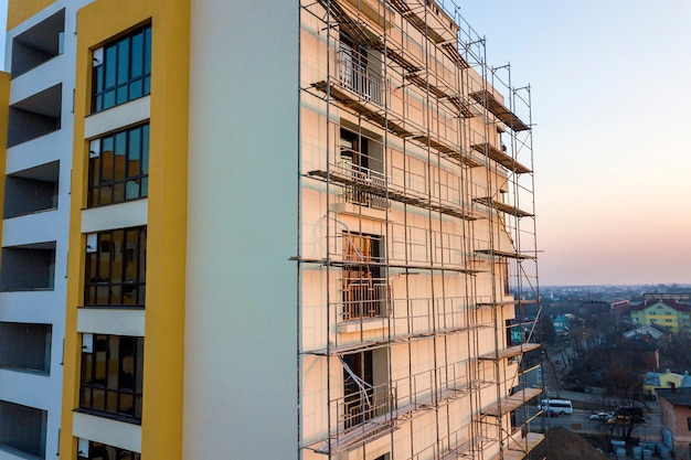 Apartment or office tall unfinished building under construction. Brick wall in scaffolding, shiny windows and tower crane on urban landscape and blue sky background. Drone aerial photography.