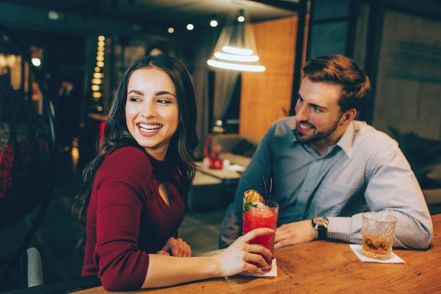 Apicture of happy girl sitting with a handsome guy in the bar. She is looking somewhere aside and smiling. Young man is looking at her and smiling as well.