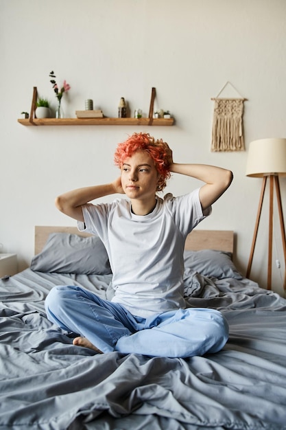 Photo appealing queer person with vibrant red hair sitting on bed and looking away leisure time