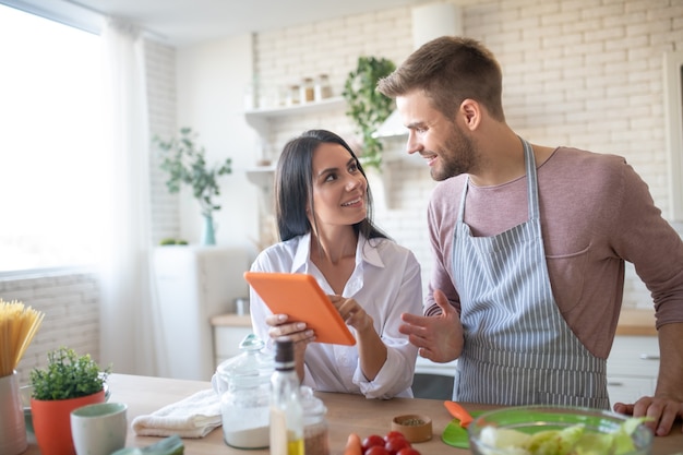 Photo appealing smiling wife holding orange tablet while cooking with husband