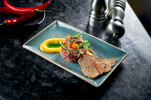 Photo an appetizer before the main course - beef steak tartare served with croutons, cappers, yolk, pickles in a blue plate on a marble table. restaurant food. raw meat