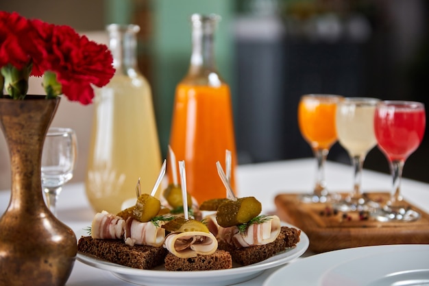 An appetizer of pickled cucumber and bacon on a piece of rye bread against the background of a served table with multicolored alcoholic drinks. Close-up, selective focus