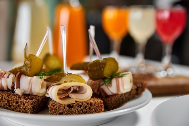 An appetizer of pickled cucumber and bacon on a piece of rye bread against the background of a served table with multicolored alcoholic drinks. Close-up, selective focus