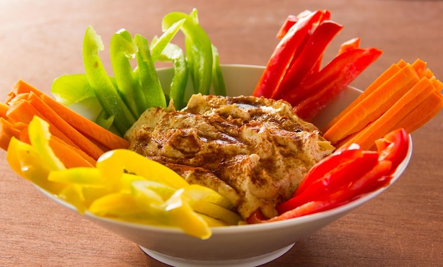 Appetizer white bowl with hummus in the center and green, red, yellow bell peppers and carrots