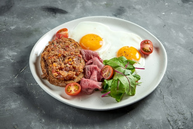 Appetizing breakfast - fried eggs with roast beef, tomatoes and salad, toast with stewed eggplant in a white plate on a gray surface