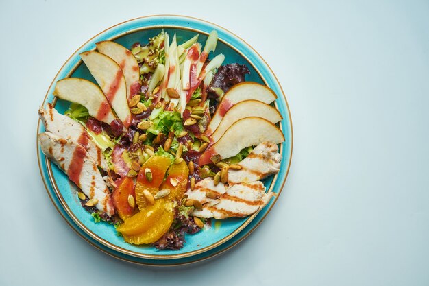 Appetizing buddha bowl with chicken, oranges, lettuce and pear in a blue ceramic plate on a gray surface. Close up view. Healthy food