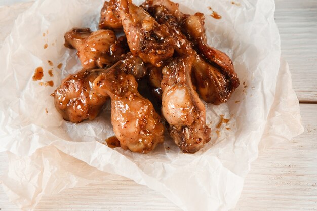 Appetizing fresh baked chicken wings in sauce, fast food. Traditional american cuisine, poultry legs in paper on white wooden table, closeup.