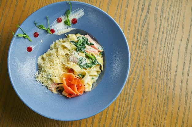 Appetizing, home-made tagliatelle pasta with spinach and salmon, parmesan in a blue bowl on a wooden surface. Italian cuisine. Add noise on post. Selective focus