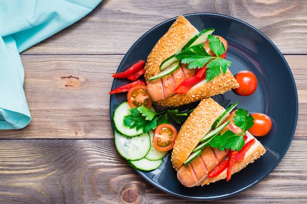 Appetizing hot dogs from fried sausages, sesame buns and fresh vegetables on a plate on a wooden table