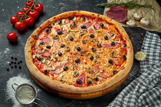 Appetizing italian pizza with cheese, ham, mushrooms, olives and red sauce in a composition with ingredients on a black table