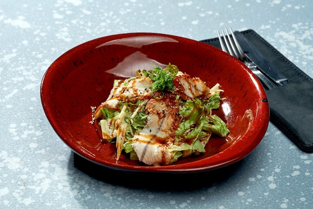 Appetizing salad with eel, teriyaki sauce, lettuce in a red plate on a blue surface