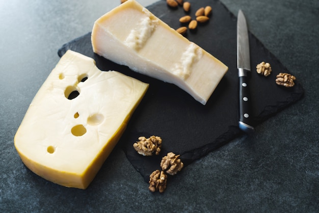 Appetizing solid cheese slices on a black plate with knife, almonds and nuts