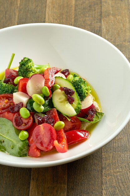 Appetizing vegetable salad with cucumber, tomatoes, radish, beans and broccoli with green butter in a white bowl on wooden table