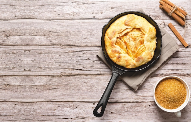 Apple galette in a black pan brown sugar in a cup and cinnamon sticks on a wooden table