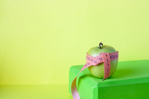 An apple girded with a centimeter tape stands on a yoga brick on a green background