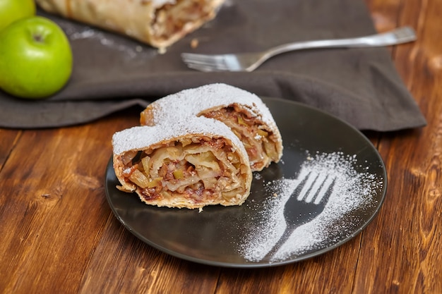 Apple strudel with icing sugar on black plate, wooden table