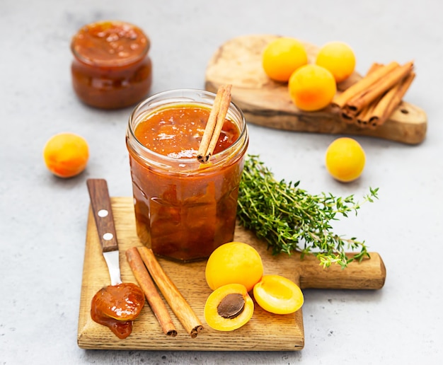 Apricot jam in a glass jar, fresh apricots, thyme and cinnamon on grey surface