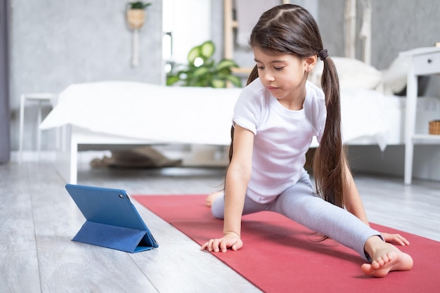 Arab kid doing stretching exercising online with tablet on fitness mat at home in modern apartment