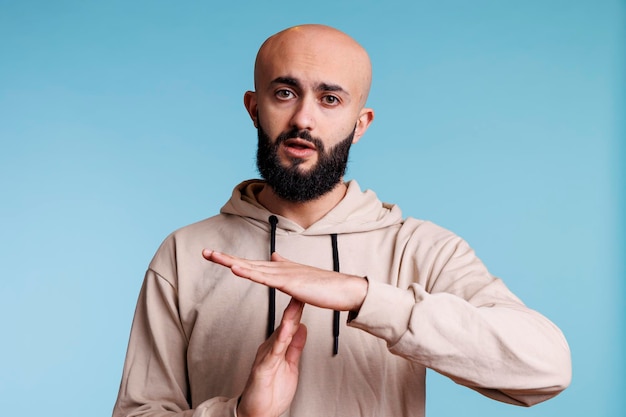 Arab man making time out gesture with hands while looking at camera. Young bald bearded person pausing conversation, showing interruption signal with arms studio portrait