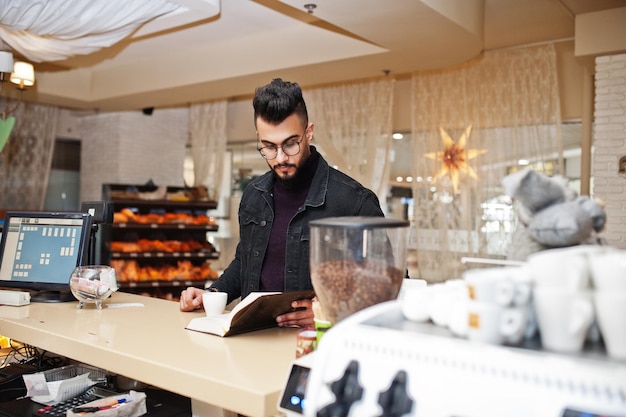 Arab man wear on black jeans jacket and eyeglasses in cafe drink coffee at bar with book Stylish and fashionable arabian model guy