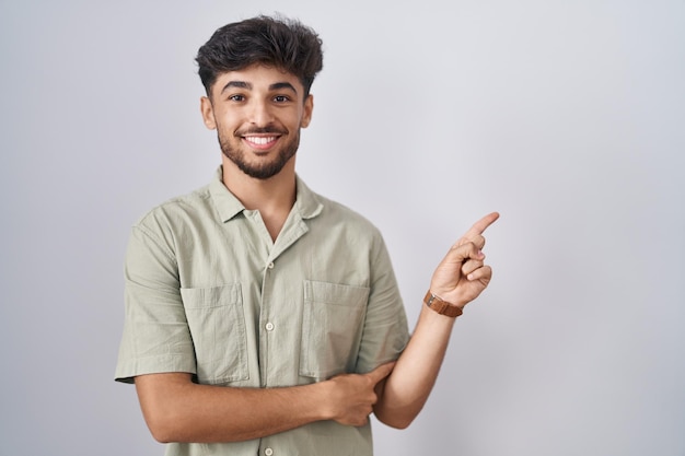 Arab man with beard standing over white background with a big smile on face, pointing with hand finger to the side looking at the camera.