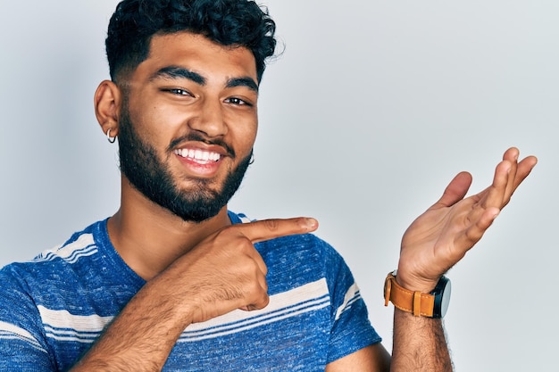 Arab man with beard wearing casual striped t shirt amazed and smiling to the camera while presenting with hand and pointing with finger.