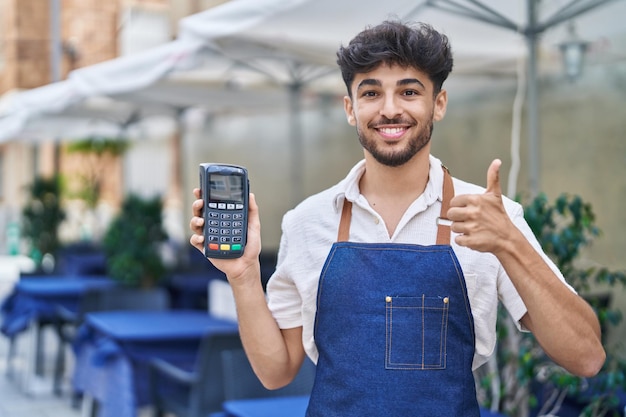 Arab man with beard wearing waiter apron at restaurant terrace holding dataphone smiling happy and positive, thumb up doing excellent and approval sign