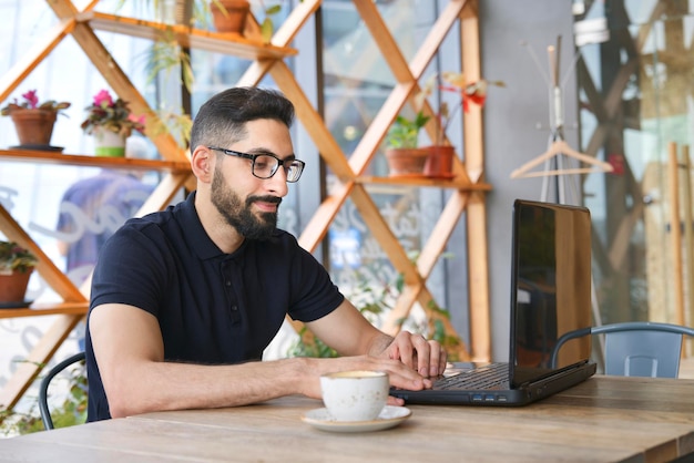 Arab Muslim ethnic guy young man freelancer sitting in cafe working on laptop drink coffee in cafe